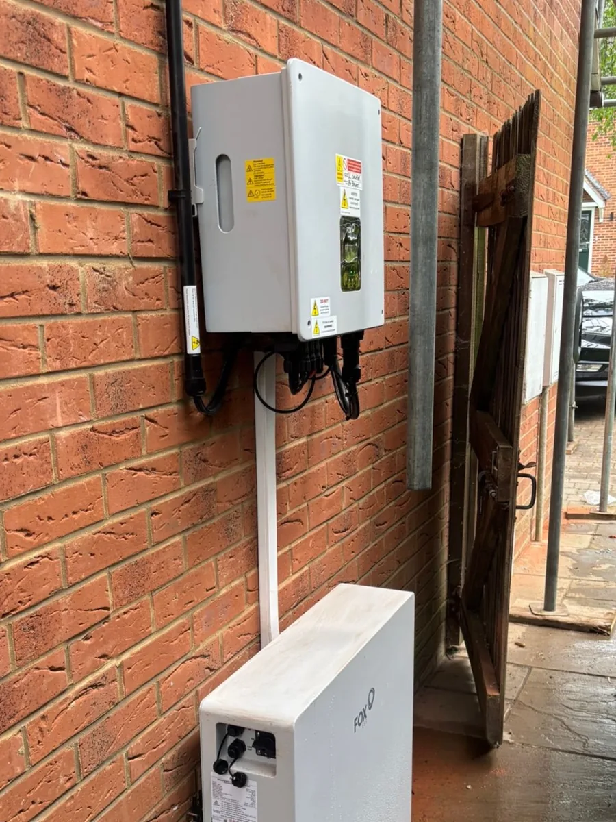 A wall mounted inverter above a white Fox ESS battery on a brick wall at the side of a house.