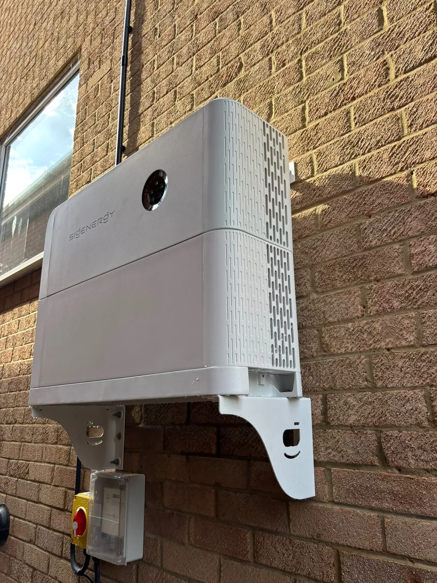 A white Sigenergy battery on a bracket on a brick wall, with an isolator switch fitted below it.